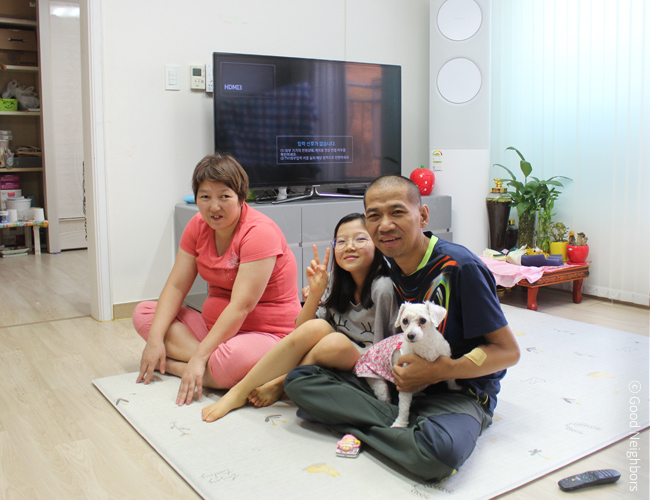 거실에서 좌측부터 어머니, 슬빈이, 아버지 그리고 하얀 강아지가 밝게 웃으며 카메라를 응시하는 모습 사진